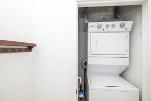 A compact laundry room with a stacked washer and dryer, a metal drying rack, and a water heater. - Walk to NoDa Arts District + Brew Pubs | King Bed (Charlotte)