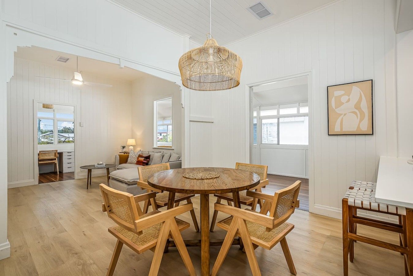 A dining area with a round wooden table and chairs, a hanging light fixture, and a view through a window.