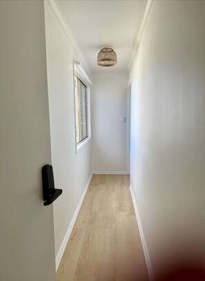A narrow hallway with white walls, a wooden floor, a door with a black handle, and a window with a white frame.