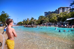 A beach with clear water, palm trees, and a building in the background.