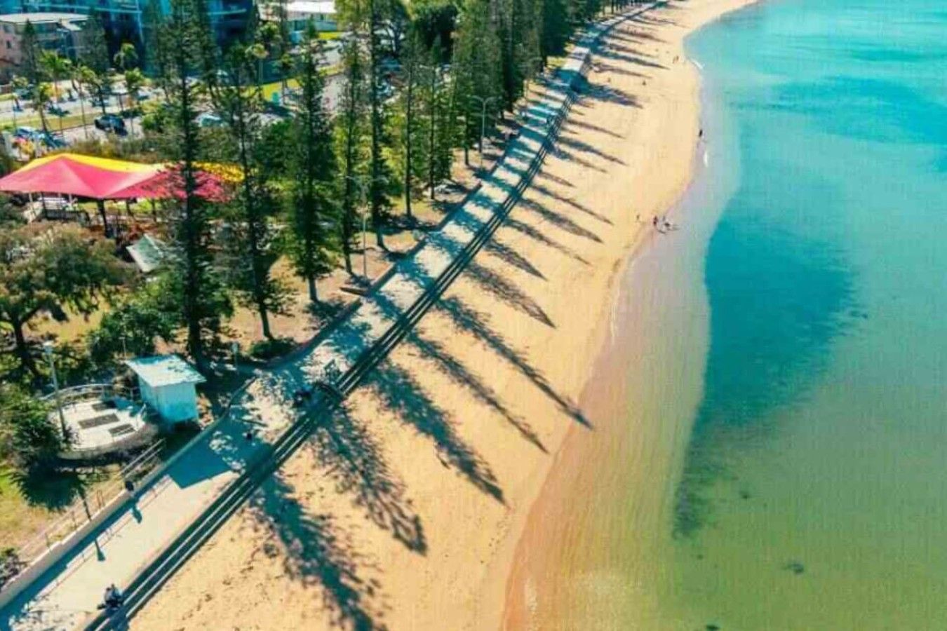 Eine Luftaufnahme eines Strandes mit einem gepflasterten Weg, Bäumen und einigen Gebäuden, darunter ein kleines Haus und ein roter Sonnenschirm.
