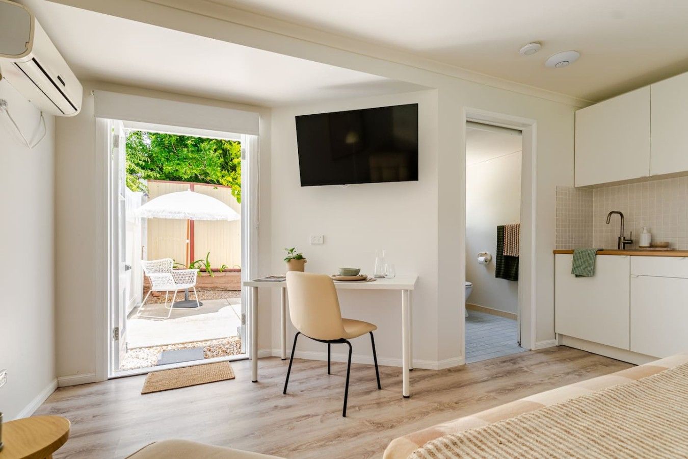 A modern, well-lit room with a dining area, a TV mounted on the wall, and a kitchen with a tiled backsplash.