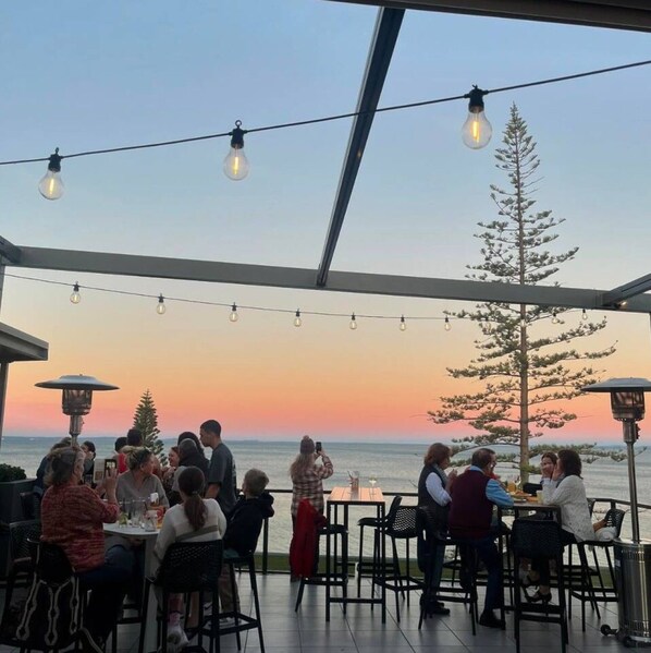 An outdoor dining area with tables and chairs, overlooking a body of water during sunset.