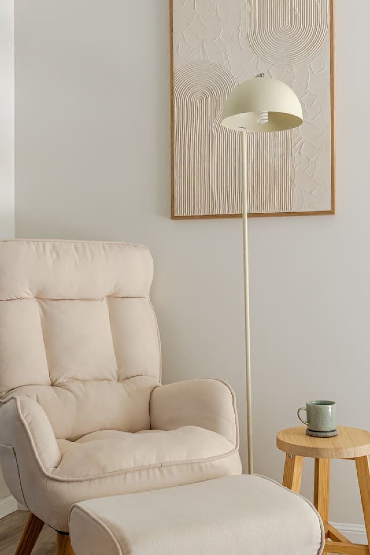 Une pièce avec un fauteuil de couleur crème, un lampadaire et une petite table en bois sur laquelle se trouve une tasse.