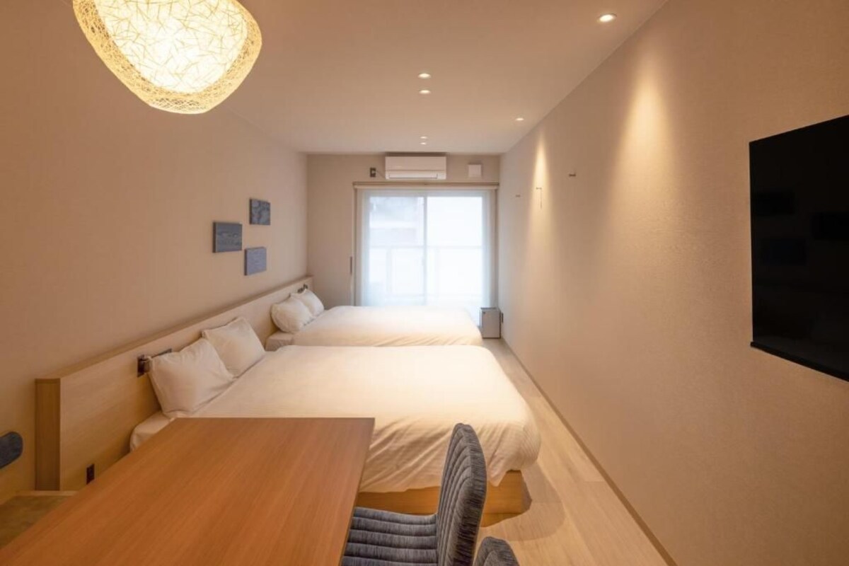 Habitació d'hotel amb dos llits, una taula de fusta, una cadira, un televisor fixat a la paret i una finestra amb llum natural.