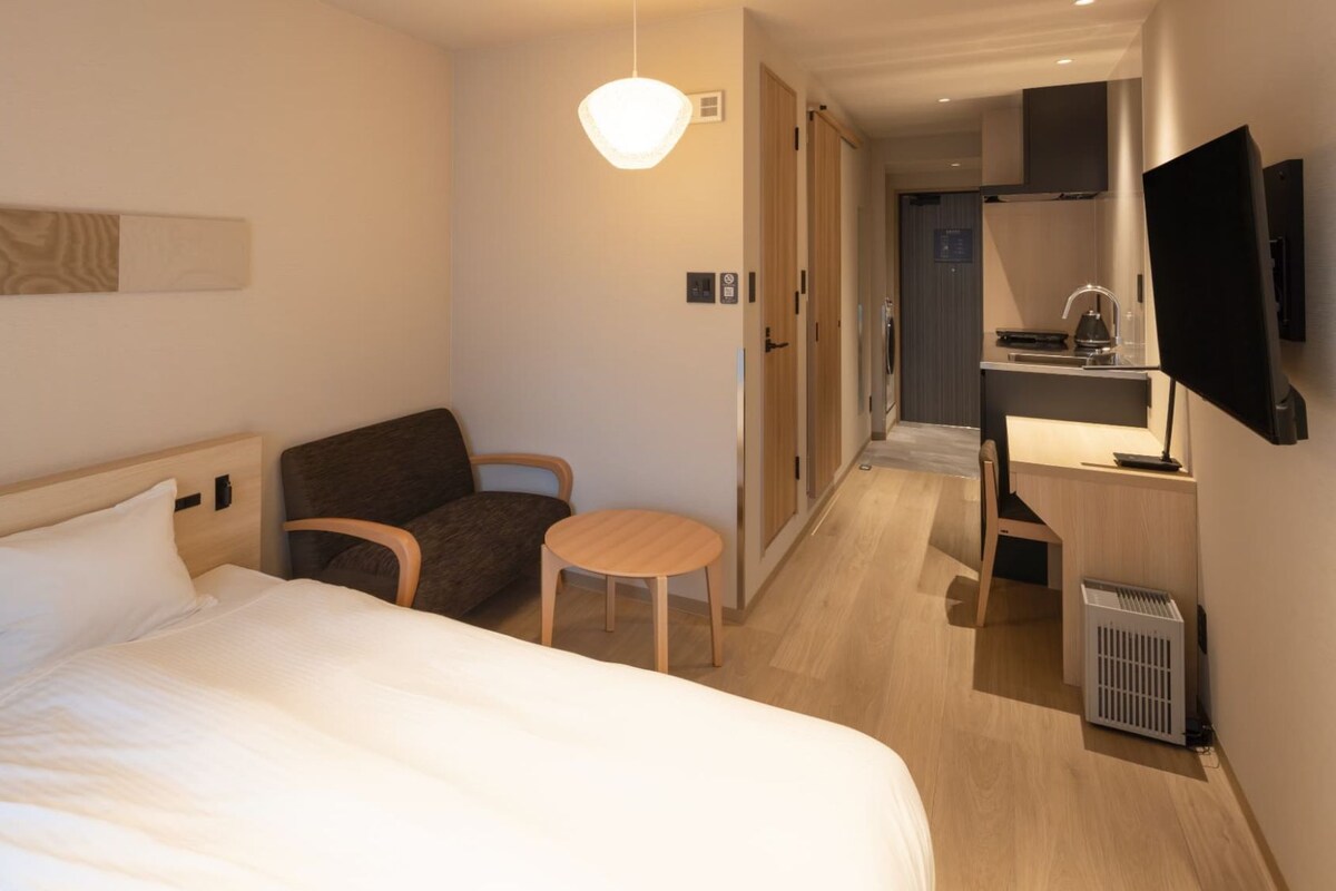 Quarto de hotel com cama, mesa pequena, cadeira, televisão e cozinha compacta.