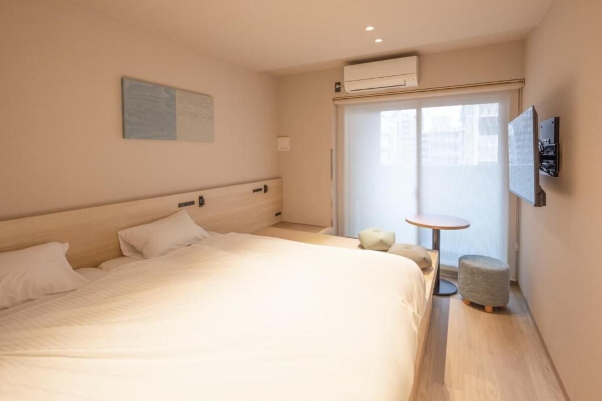 Habitació d'hotel amb un llit, una taula petita, una cadira i una finestra amb persianes.