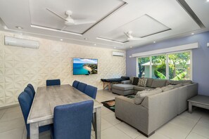 A modern living room with a dining area, a large flat-screen TV mounted on the wall, ceiling fans, and a large window with a view of greenery. - Marvelous 4-bedroom house with AC in peaceful Sosúa neighborhood (Sosúa)
