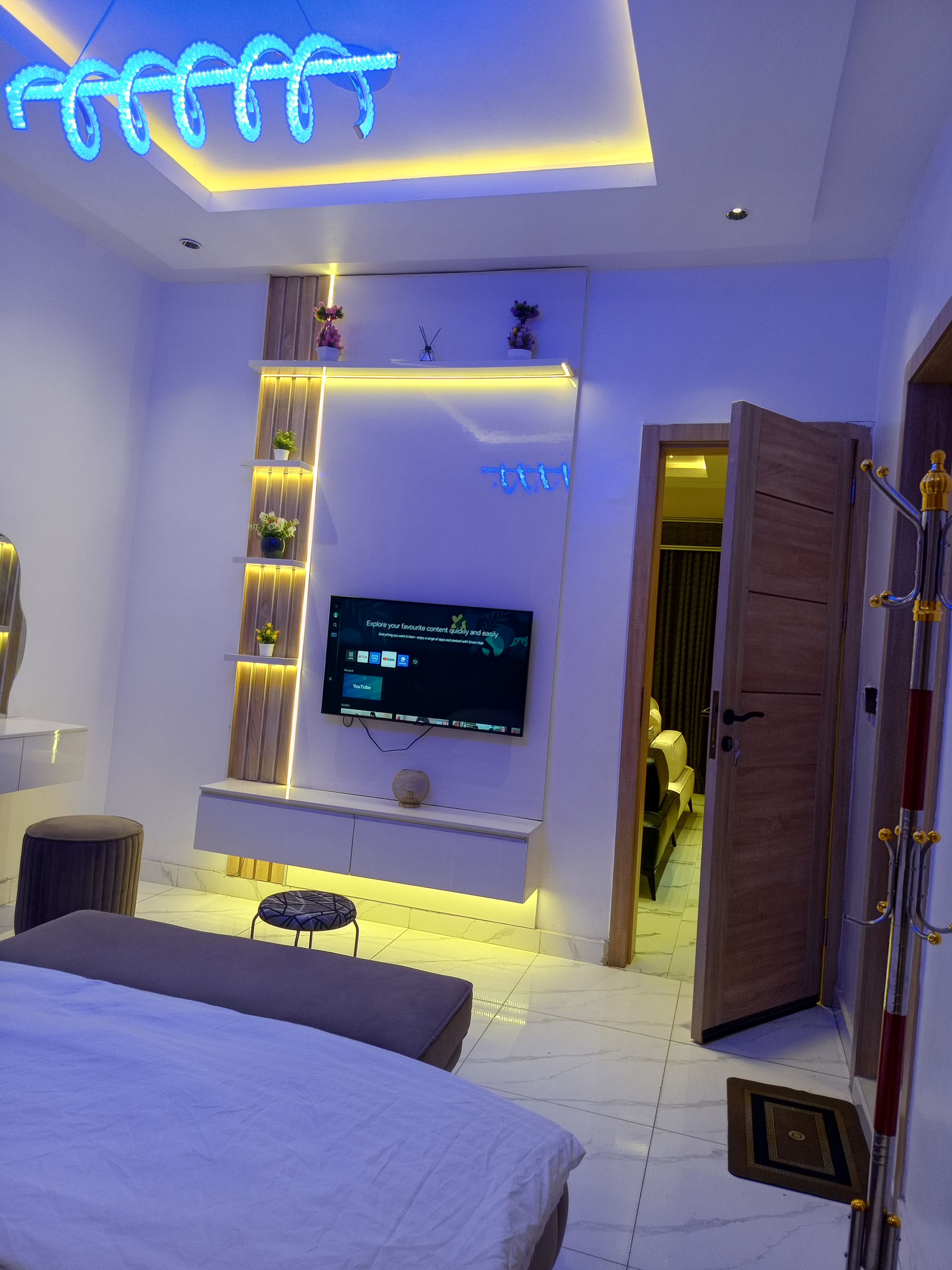 A modern bedroom with a flat-screen TV mounted on the wall, a bed, a bedside table, and a door leading to another room.