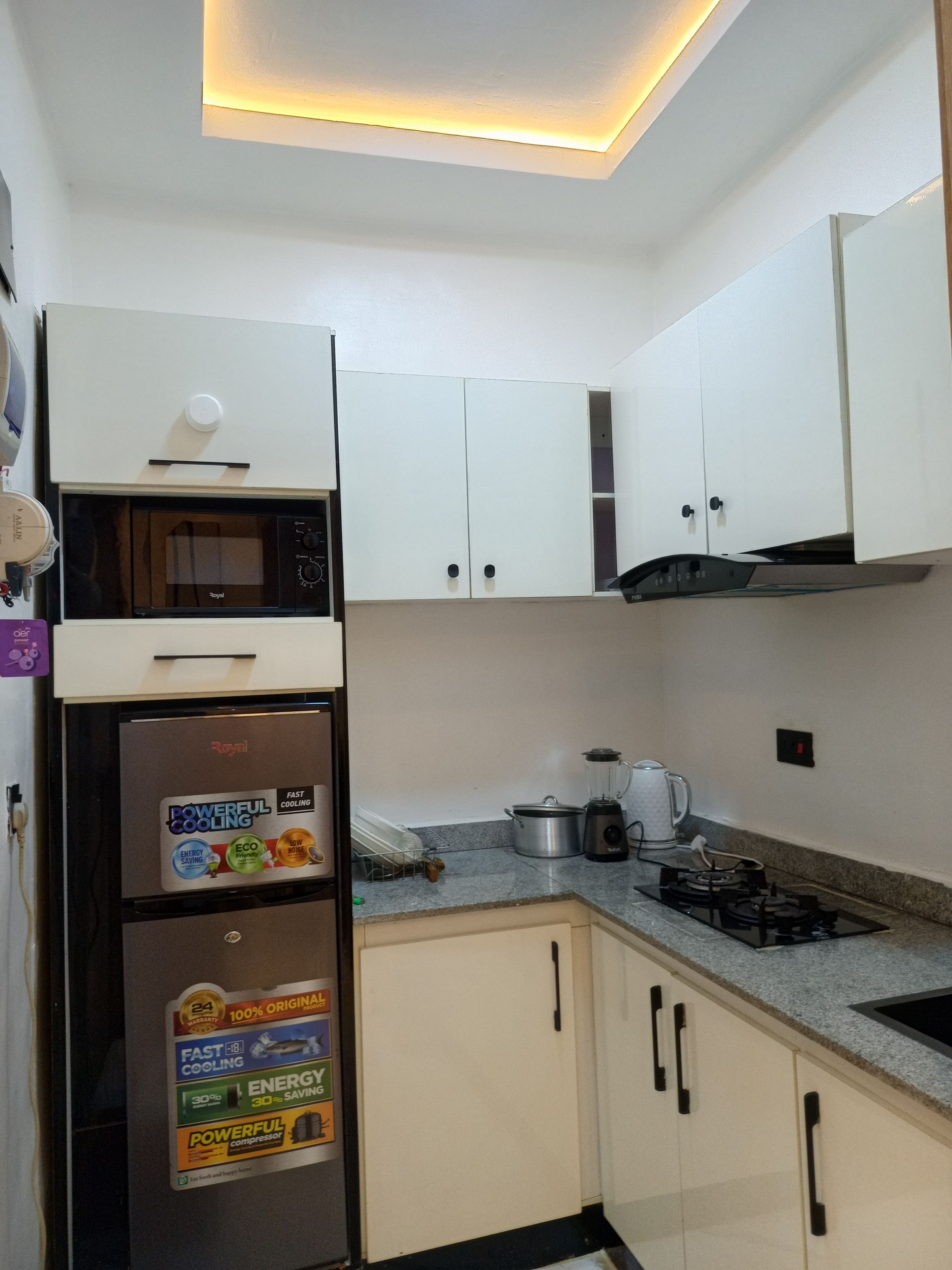 Une cuisine compacte avec des meubles blancs, un four à micro-ondes encastré, un réfrigérateur doté d’autocollants indiquant son efficacité énergétique, une cuisinière à gaz et une machine à café.