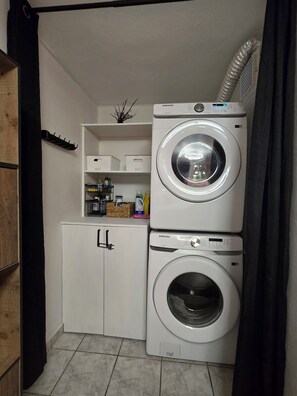 Un espace buanderie comprenant deux machines à laver superposées, un meuble avec une poignée et une étagère contenant divers objets.