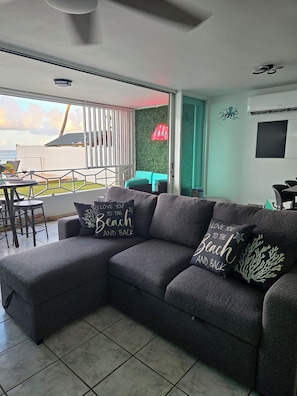 A modern living room with a grey sectional sofa, two decorative pillows, a dining area with a table and chairs, and a view of the beach through large windows. - Unforgettable, stunning Oceanfront modern condo! (Luquillo)