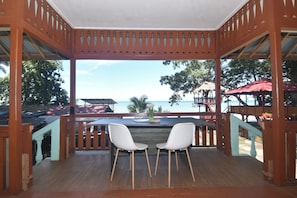 A wooden deck with two white chairs, overlooking a beach with a thatched-roof structure.