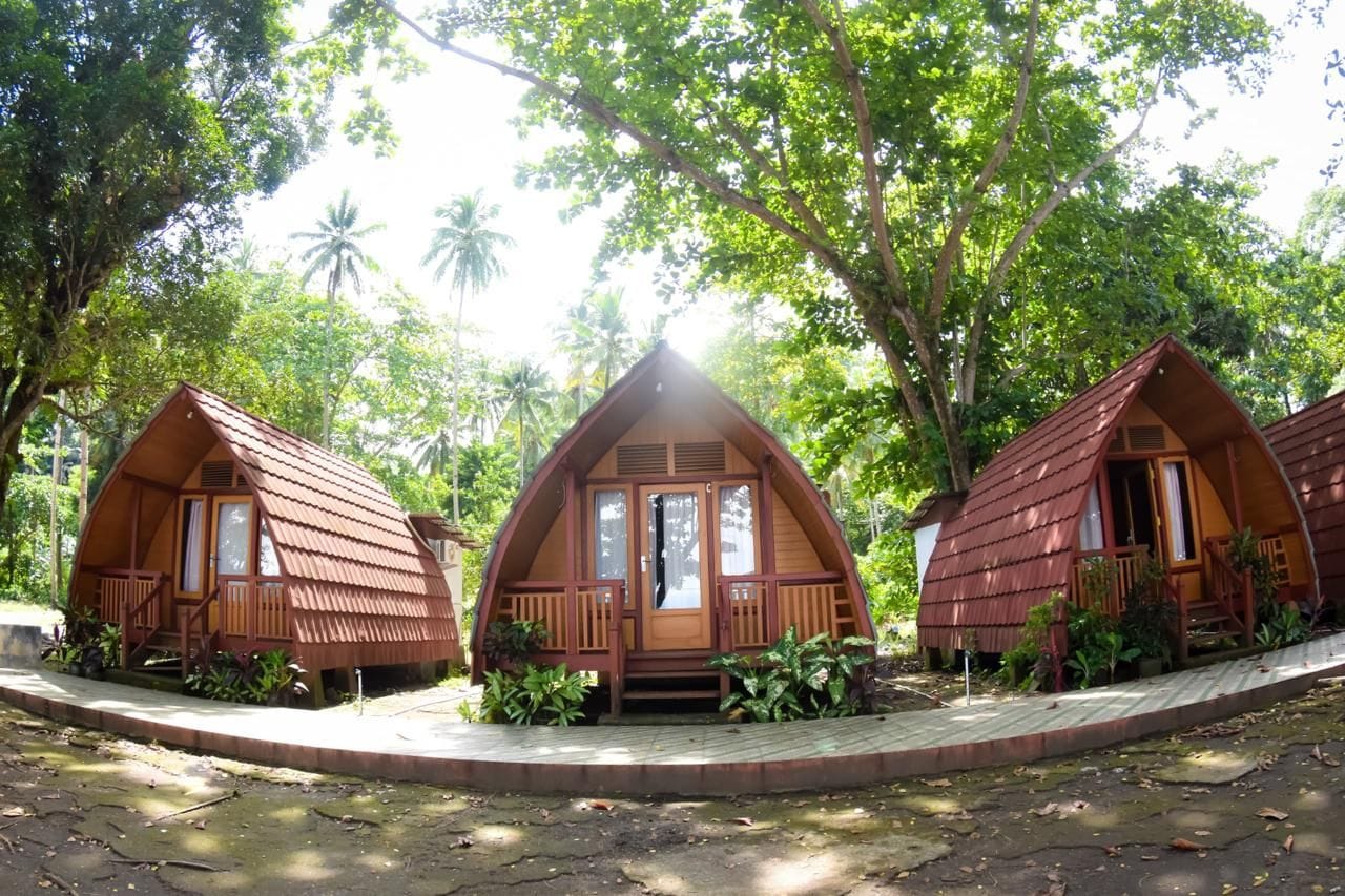 Tres cabañas de madera con techos a dos aguas, rodeadas de abundante follaje y palmeras.