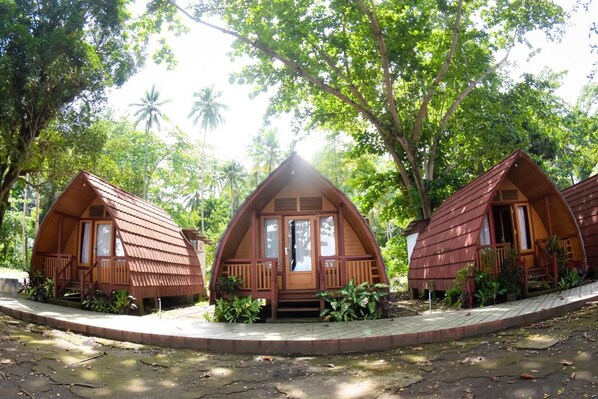 Three wooden cabins with pitched roofs, surrounded by lush greenery and palm trees.