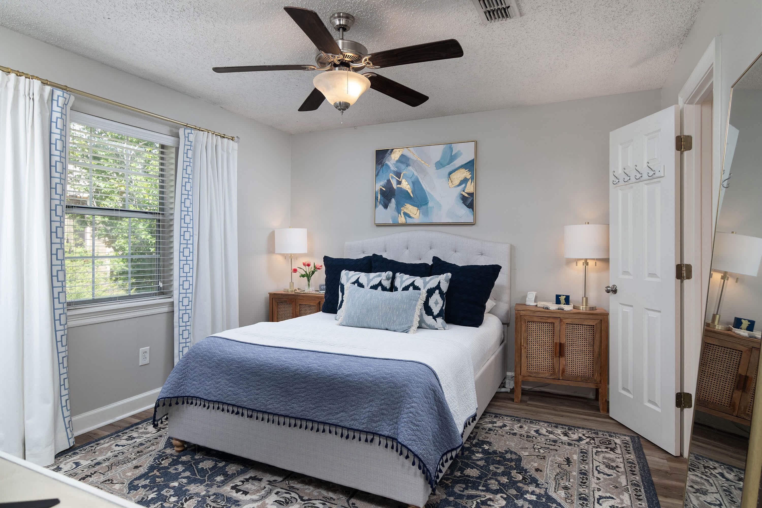 A bedroom with a bed, a ceiling fan, a window with curtains, a painting on the wall, and two bedside tables with lamps.