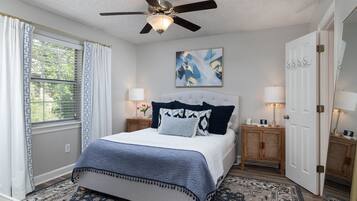 A bedroom with a bed, a ceiling fan, a window with curtains, a painting on the wall, and two bedside tables with lamps.
