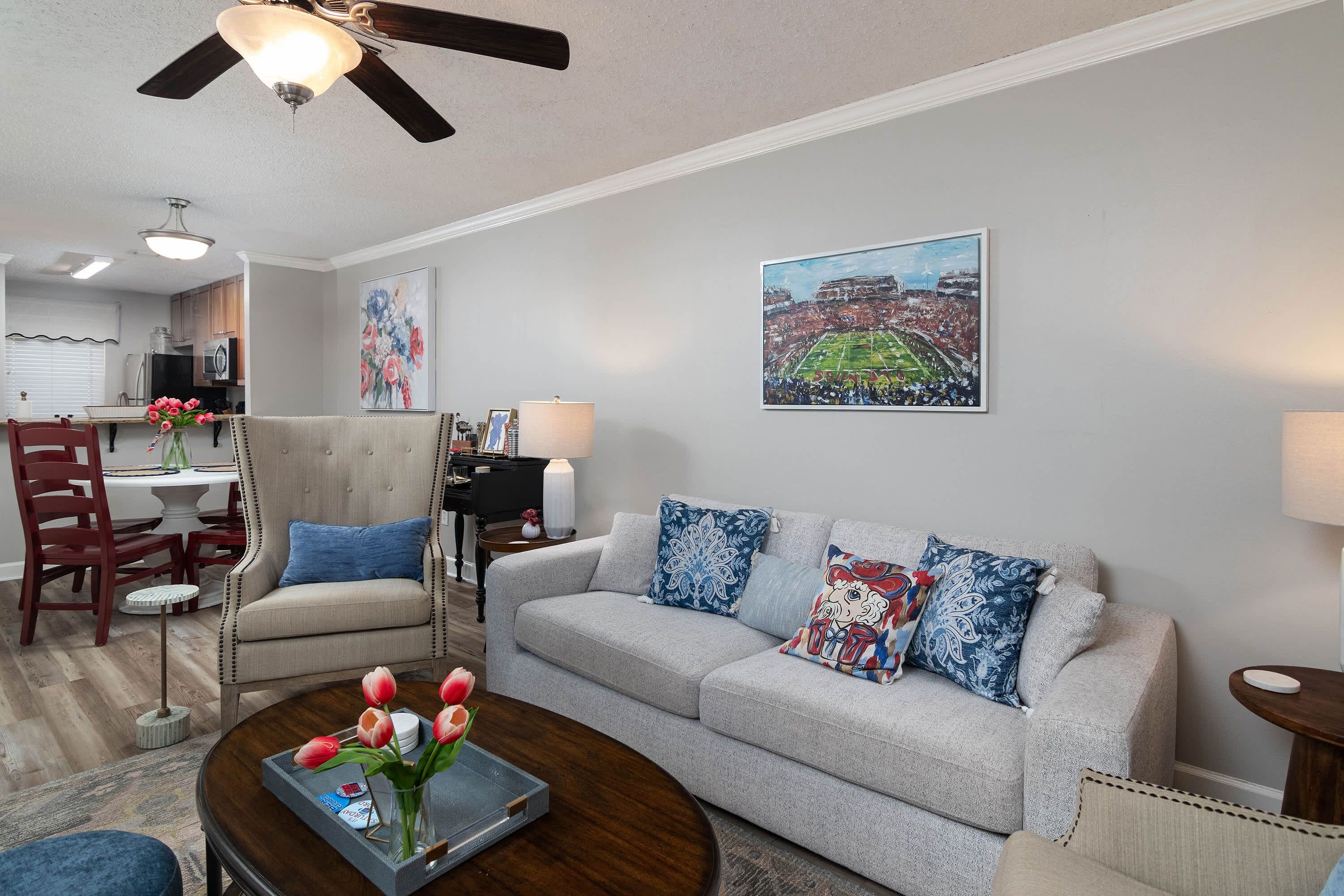 A living room with a sofa, a coffee table with tulips, a dining area with a piano, and a ceiling fan.