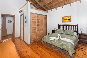 Ein Schlafzimmer mit Holzboden, einem Bett mit einer blumigen Bettdecke in Grün, einem Nachttisch mit Lampe und einem Sonnenuntergangsbild an der Wand.