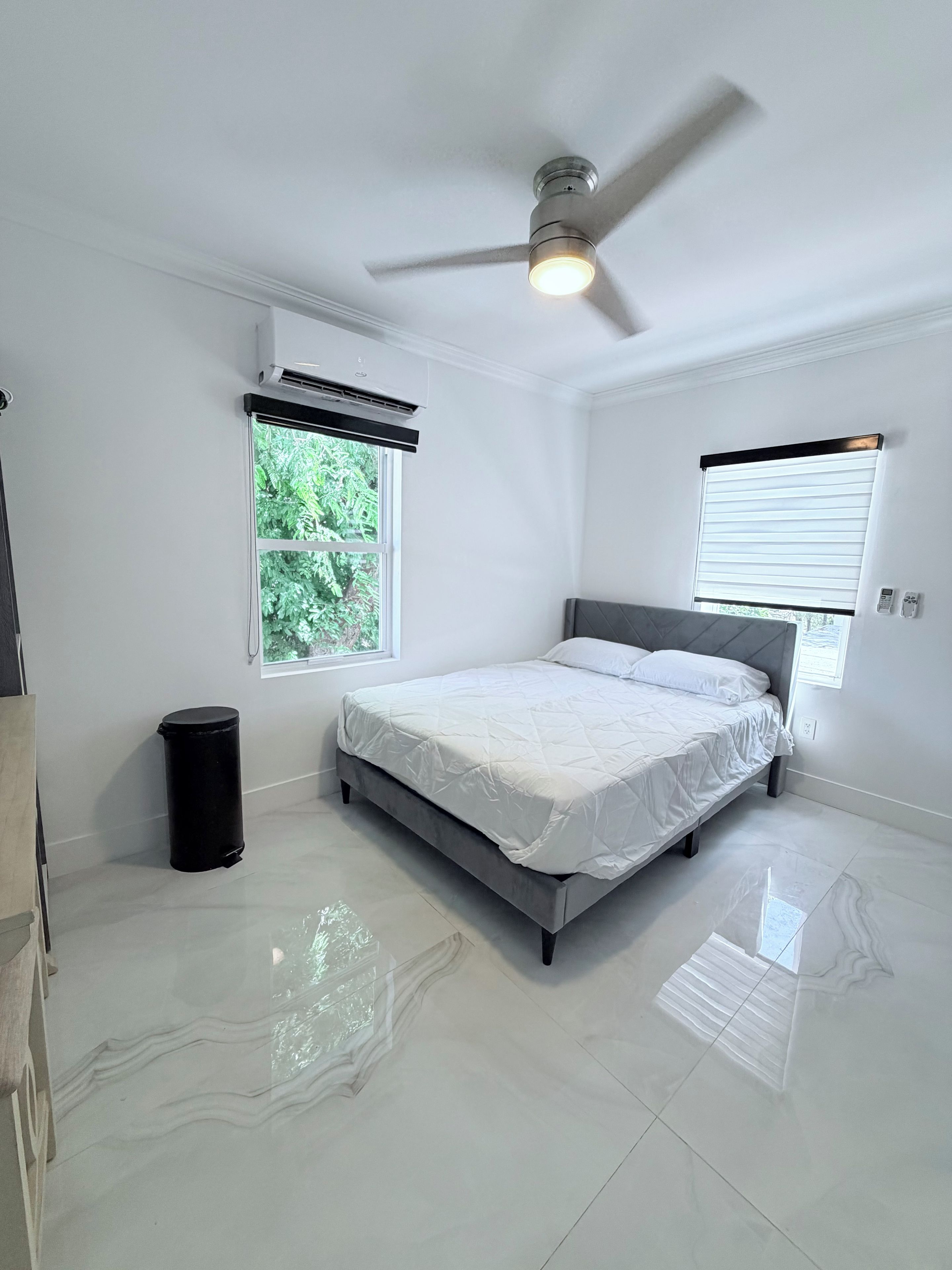 Ein Schlafzimmer mit Bett, Deckenventilator, Klimaanlage, Fenstern und einem Mülleimer.