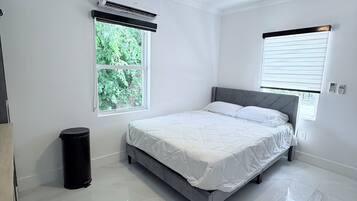 Ein Schlafzimmer mit Bett, Deckenventilator, Klimaanlage, Fenstern und einem Mülleimer.