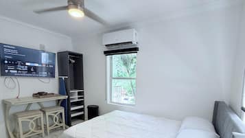 A bedroom with a bed, a ceiling fan, an air conditioning unit, a TV mounted on the wall, and a window with a view of greenery outside.