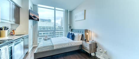 Un dormitorio con cama, mesita de noche, televisor, ventana y una vista a la ciudad, además de una kitchenette.