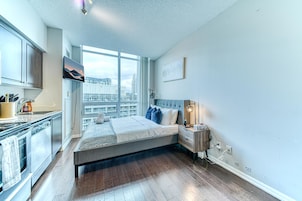 Een slaapkamer met een bed, nachtkastje, televisie, raam met stadszicht en een kitchenette.