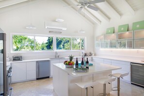A modern kitchen with a large island, bar stools, a ceiling fan, and a view of greenery through the windows. - Tropical Retreat 3-bedroom house in Long Bay Hills (Long Bay Hills)
