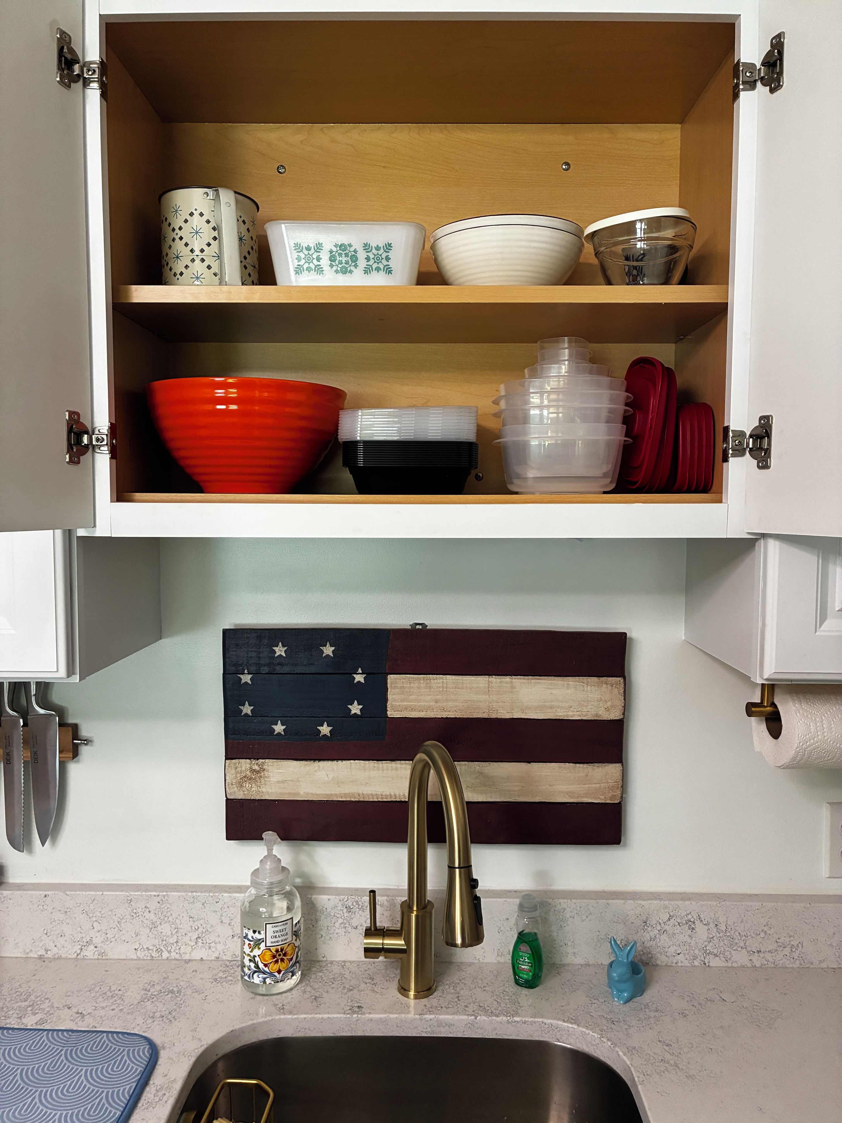 Una cucina con uno scaffale di legno sopra un lavello, su cui sono esposti utensili da cucina e una bandiera americana decorativa.