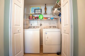 A laundry room with two front-loading washers and dryers, a shelf with cleaning supplies, and a framed picture on the wall.