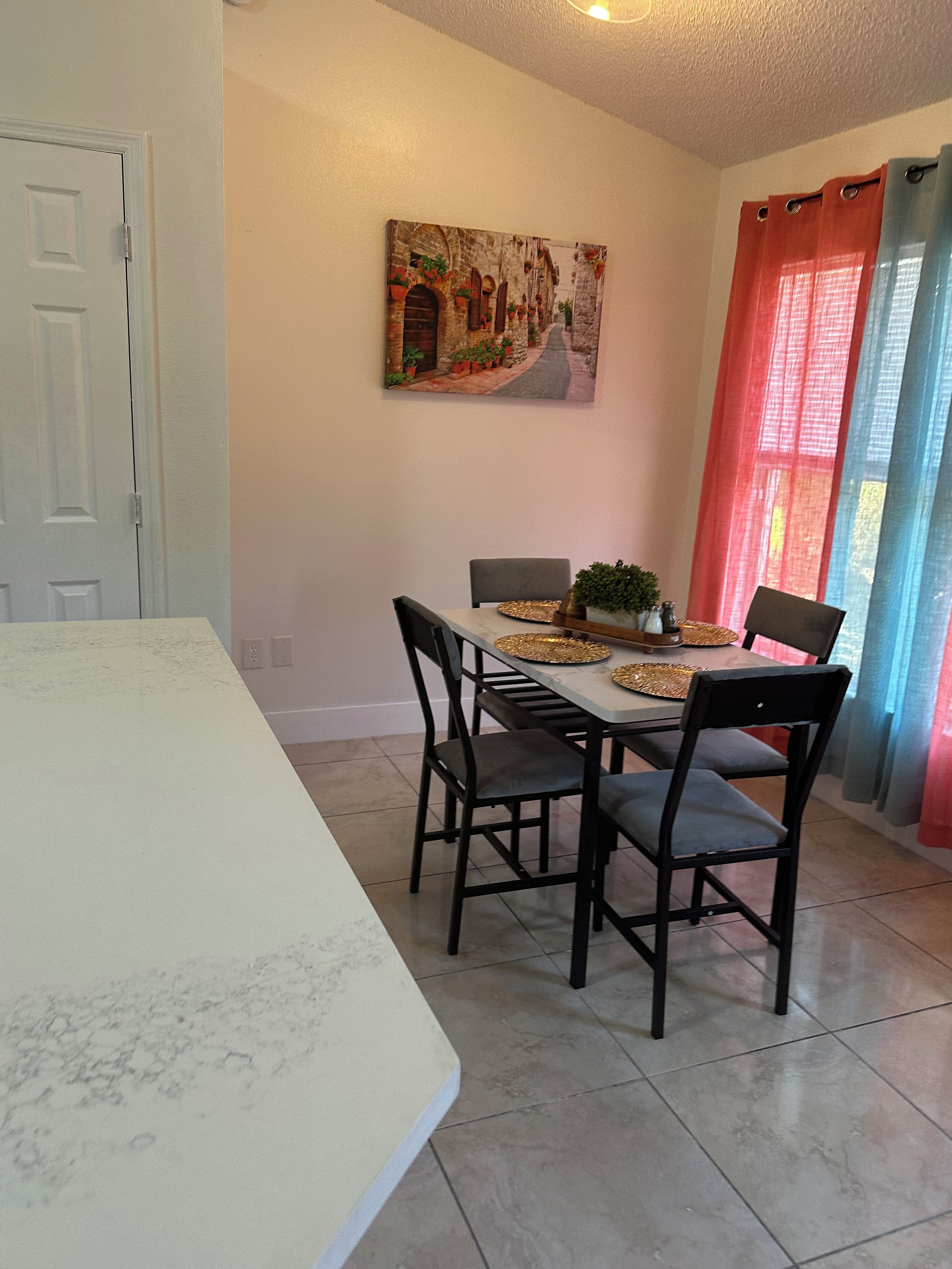 A dining area with a table set for four, chairs, a painting on the wall, and a door.
