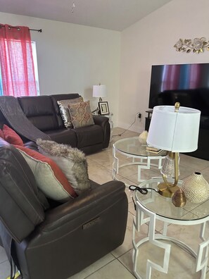 A living room with a sofa, a glass coffee table, a lamp, and a flat-screen TV. - Sunny Palm Bay Getaway  3 Bedrooms, 2 Baths & Air Conditioning. (Palm Bay)