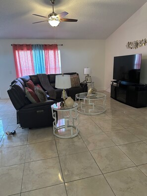 A living room with a sofa, two glass tables, a ceiling fan, a television, and a window with curtains. - Sunny Palm Bay Getaway  3 Bedrooms, 2 Baths & Air Conditioning. (Palm Bay)