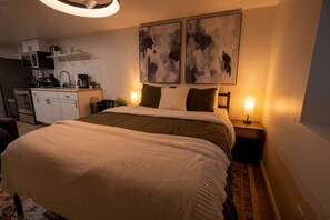 A bedroom with a bed, bedside tables, a nightstand, a wall-mounted artwork, and a kitchenette in the background. - Modern Studio - Full Kitchen (Grandview)