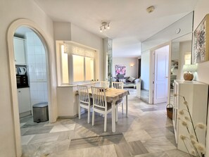 A modern kitchen with a dining area, featuring a white table and chairs, a window with a decorative valance, and a kitchen counter with appliances. - SR Cartuja 22, 2-Bedroom Apartment with Ocean View in Mijas Costa (Mijas)