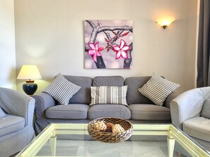 A living room with a grey sofa, patterned cushions, a glass coffee table, a wall-mounted lamp, and a framed artwork of pink flowers. - SR Cartuja 22, 2-Bedroom Apartment with Ocean View in Mijas Costa (Mijas)