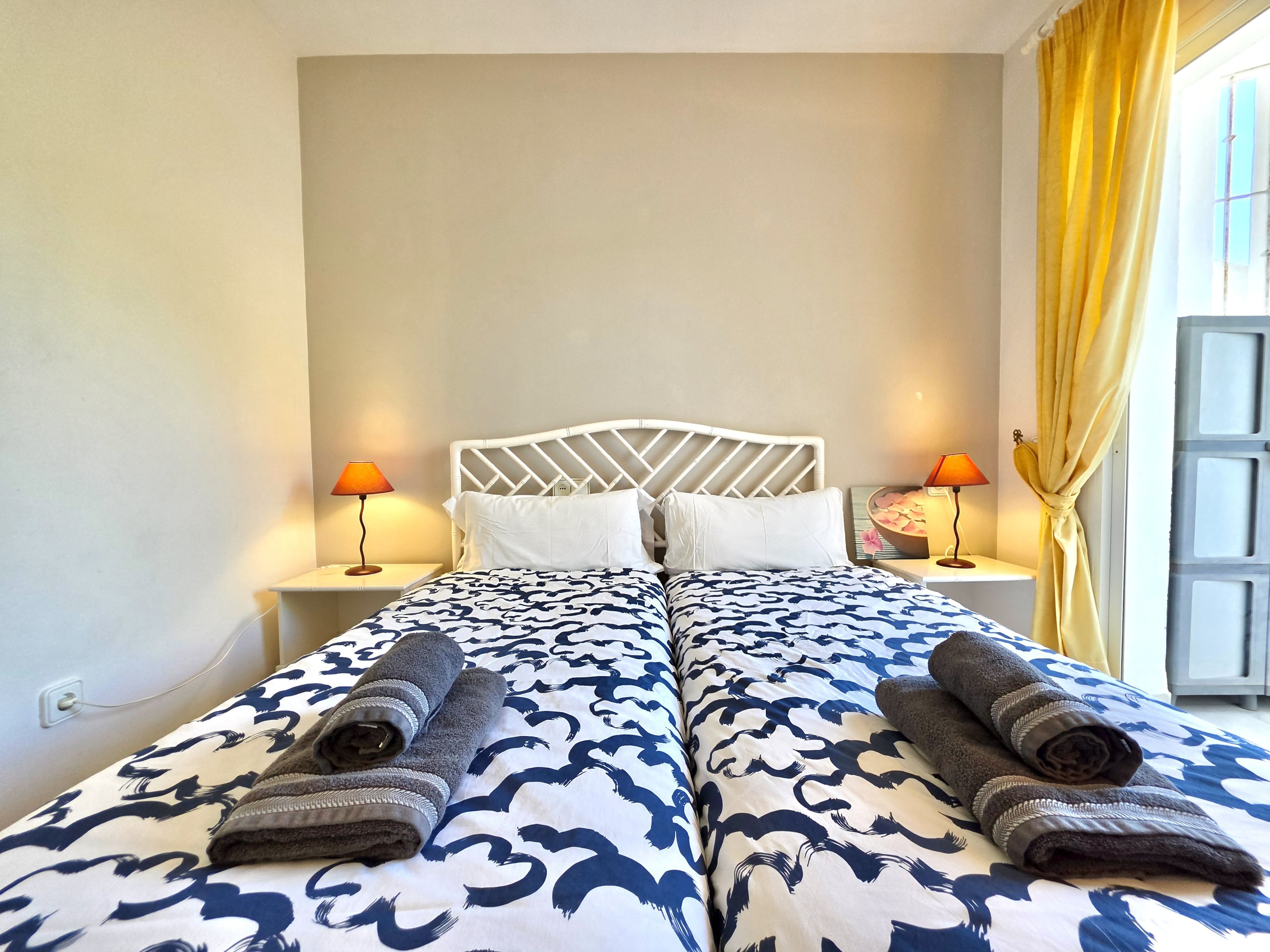 A double bed with a patterned bedspread, two bedside lamps, and a window with curtains.