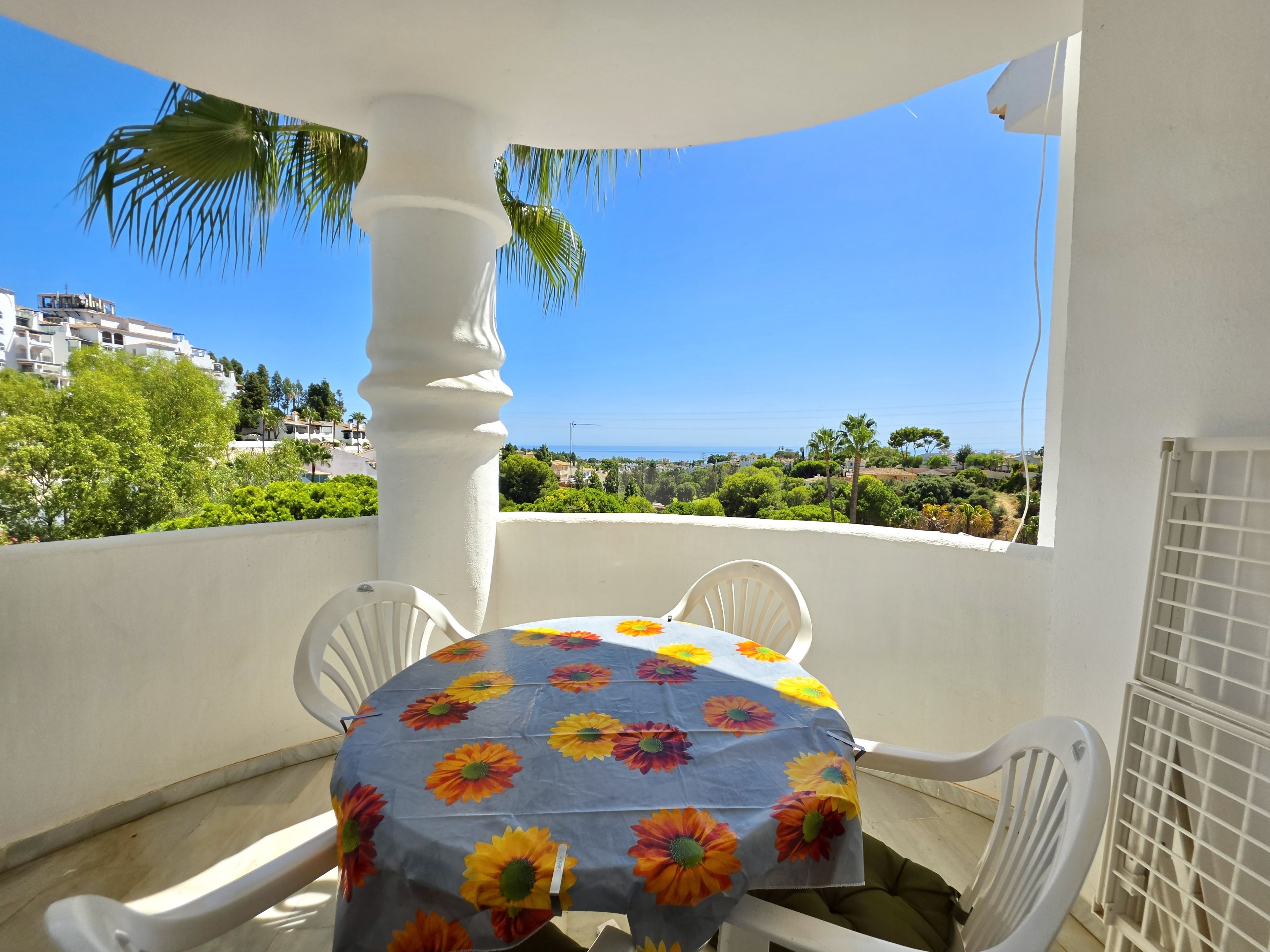 Un balcon avec une table recouverte d’une nappe à motifs floraux, des chaises blanches et une vue sur la mer et la verdure.