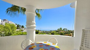 Ein Balkon mit Tisch, bedeckt mit einer blumigen Tischdecke, weißen Stühlen und Blick auf das Meer und Grün.