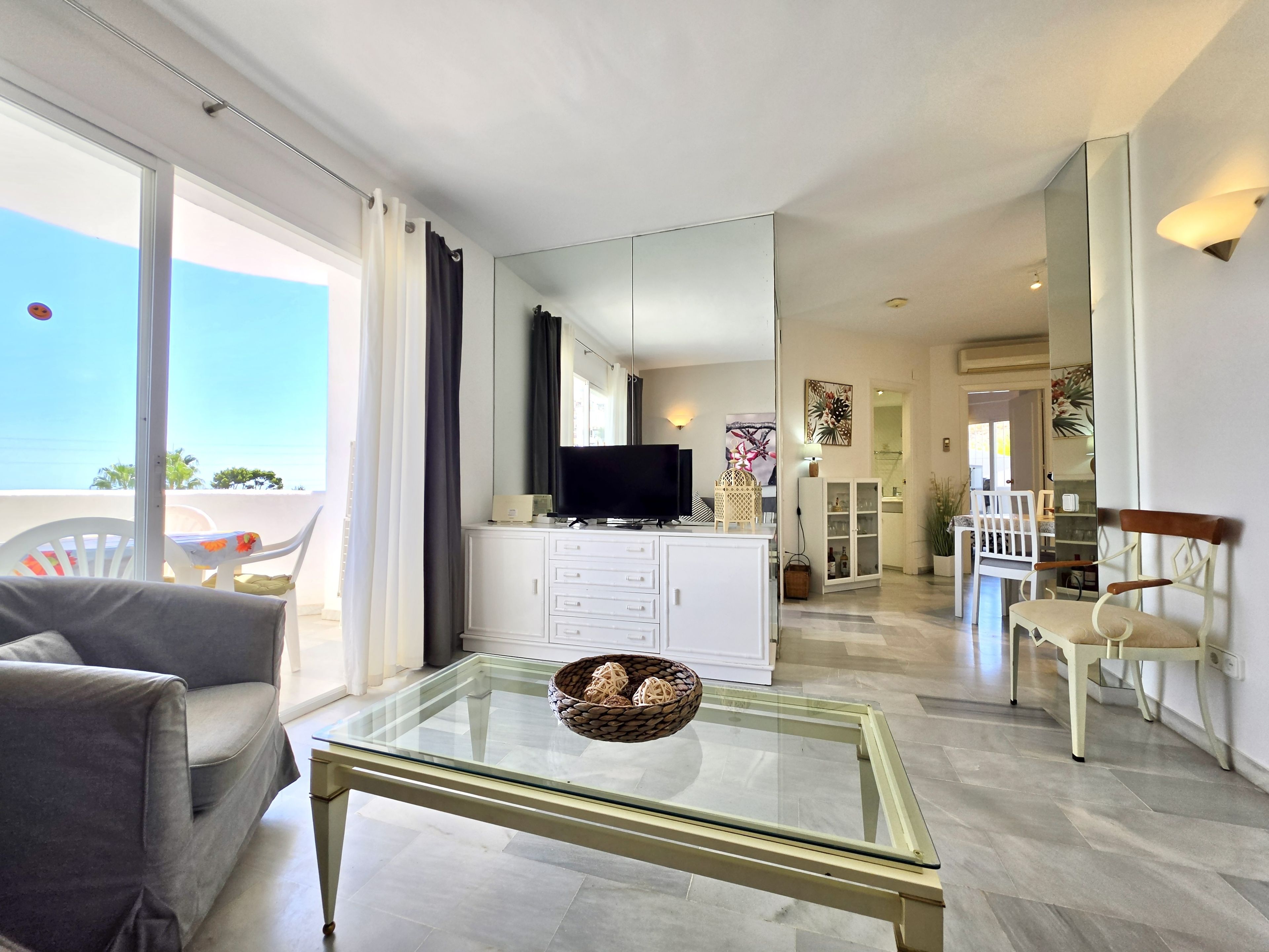 Una sala moderna con una mesa de centro de vidrio, un sofá, un televisor y vistas al exterior.