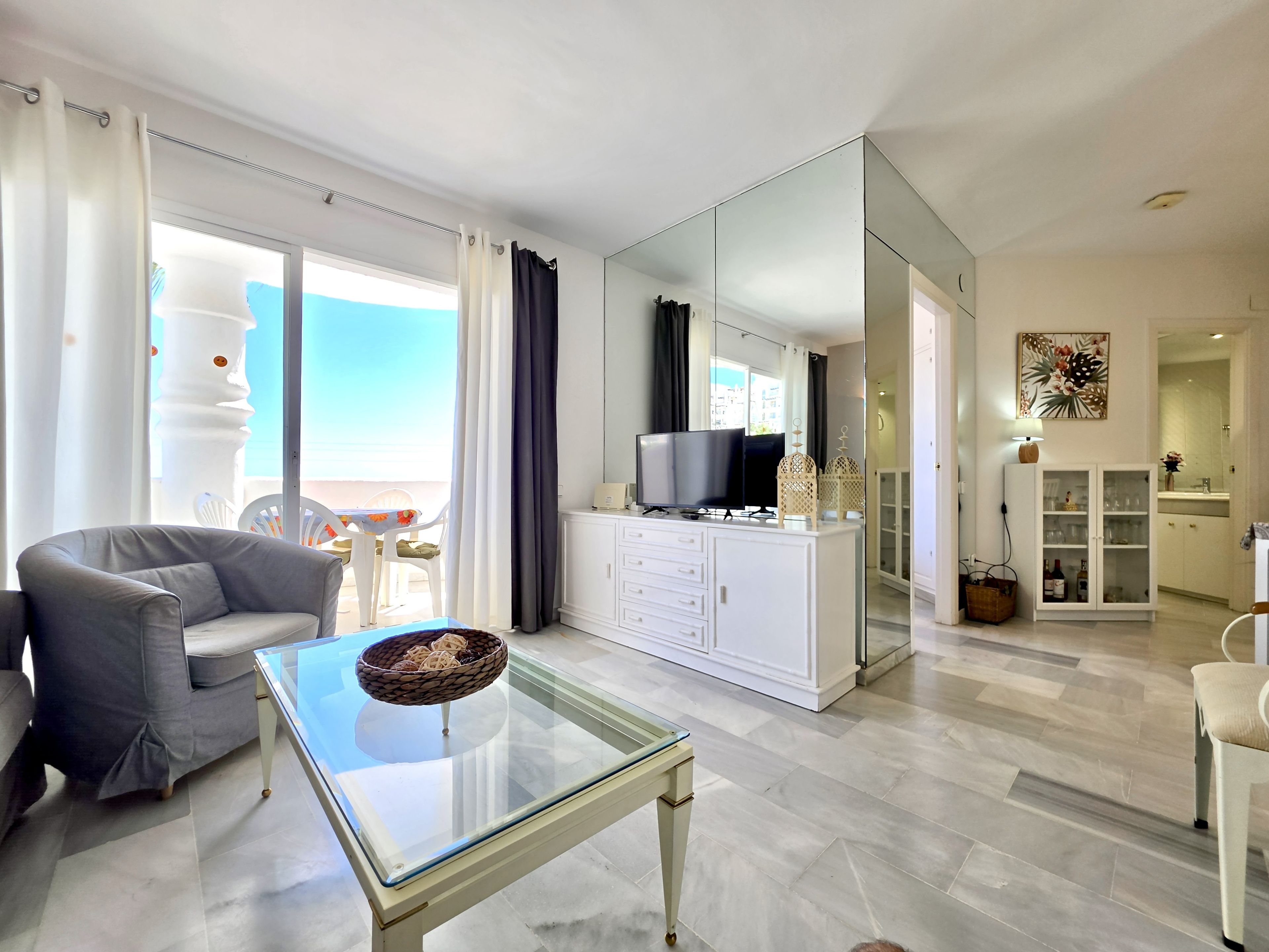 A modern living room with a glass coffee table, a grey sofa, a television, and a view of the outdoors.