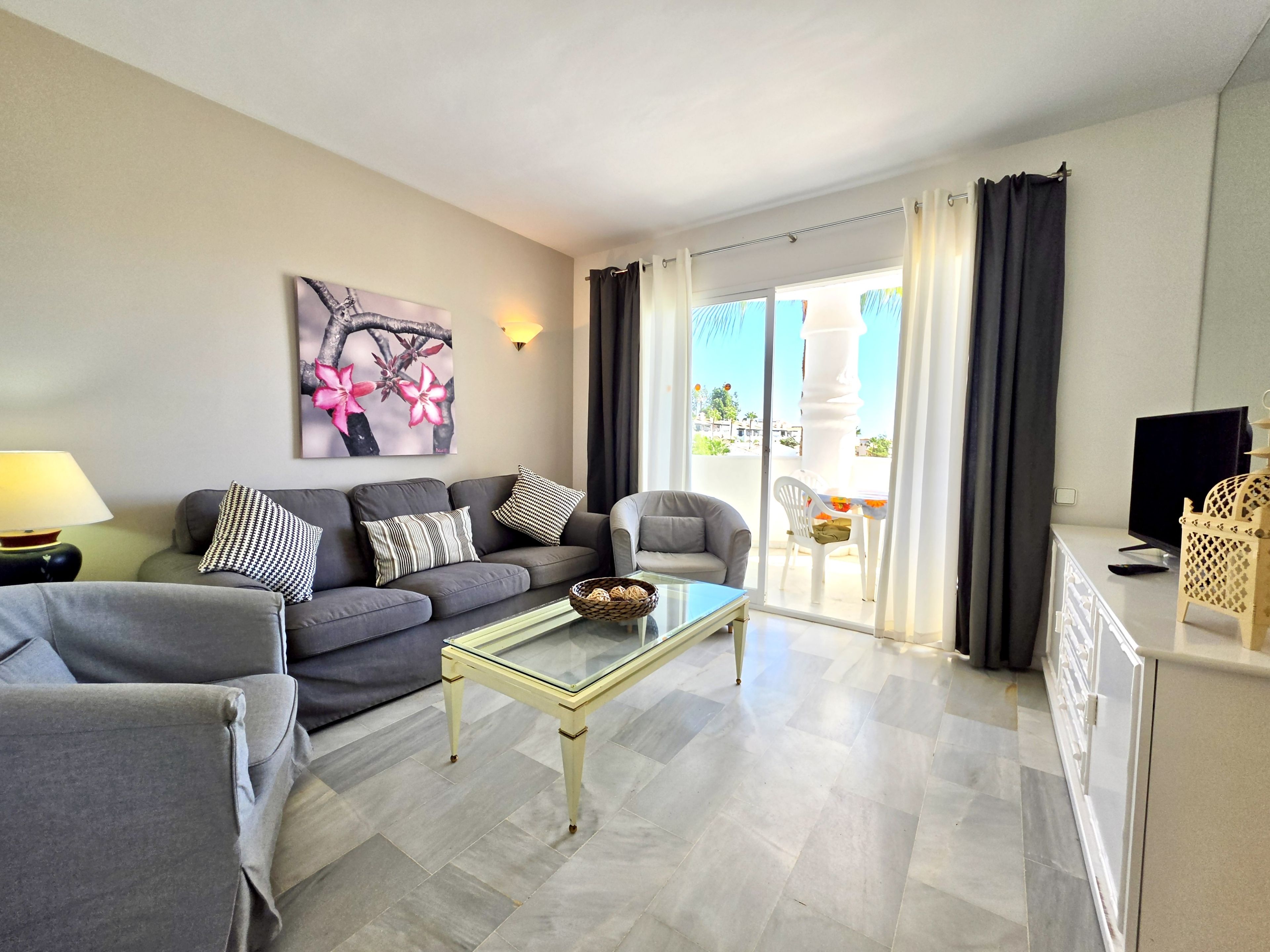 Ein Wohnzimmer mit Sofa, Sesseln, Couchtisch, Fernseher und einem Balkon mit Vorhängen.