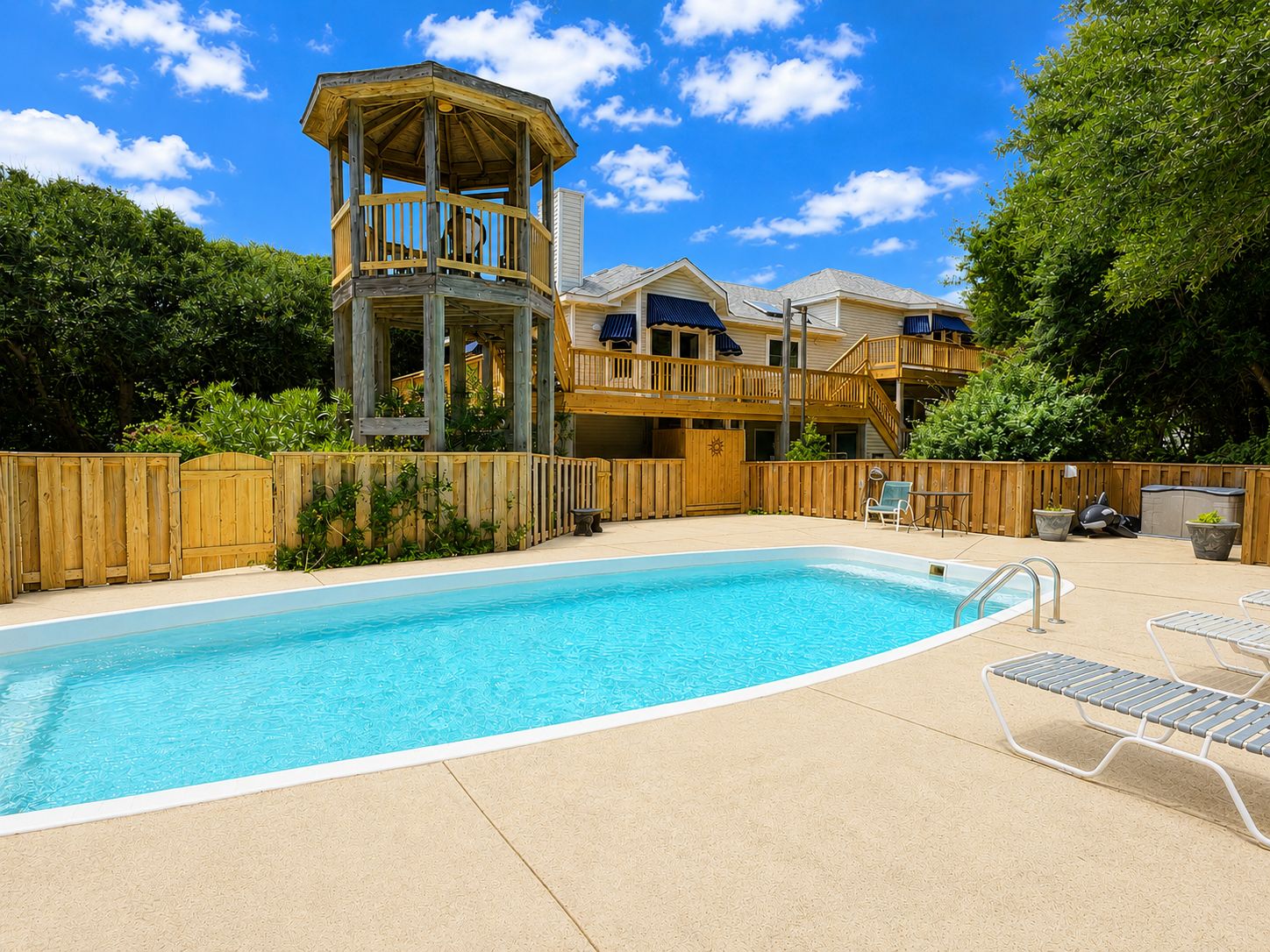 A swimming pool with a wooden deck, a wooden decked structure, and a fenced area with a view of a multi-story building.