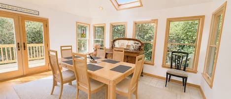 A dining area with a table and chairs, a kitchen in the background, and large windows.