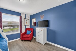 Une chambre à coucher avec un fauteuil rouge, une commode blanche, un téléviseur et une fenêtre avec des rideaux.