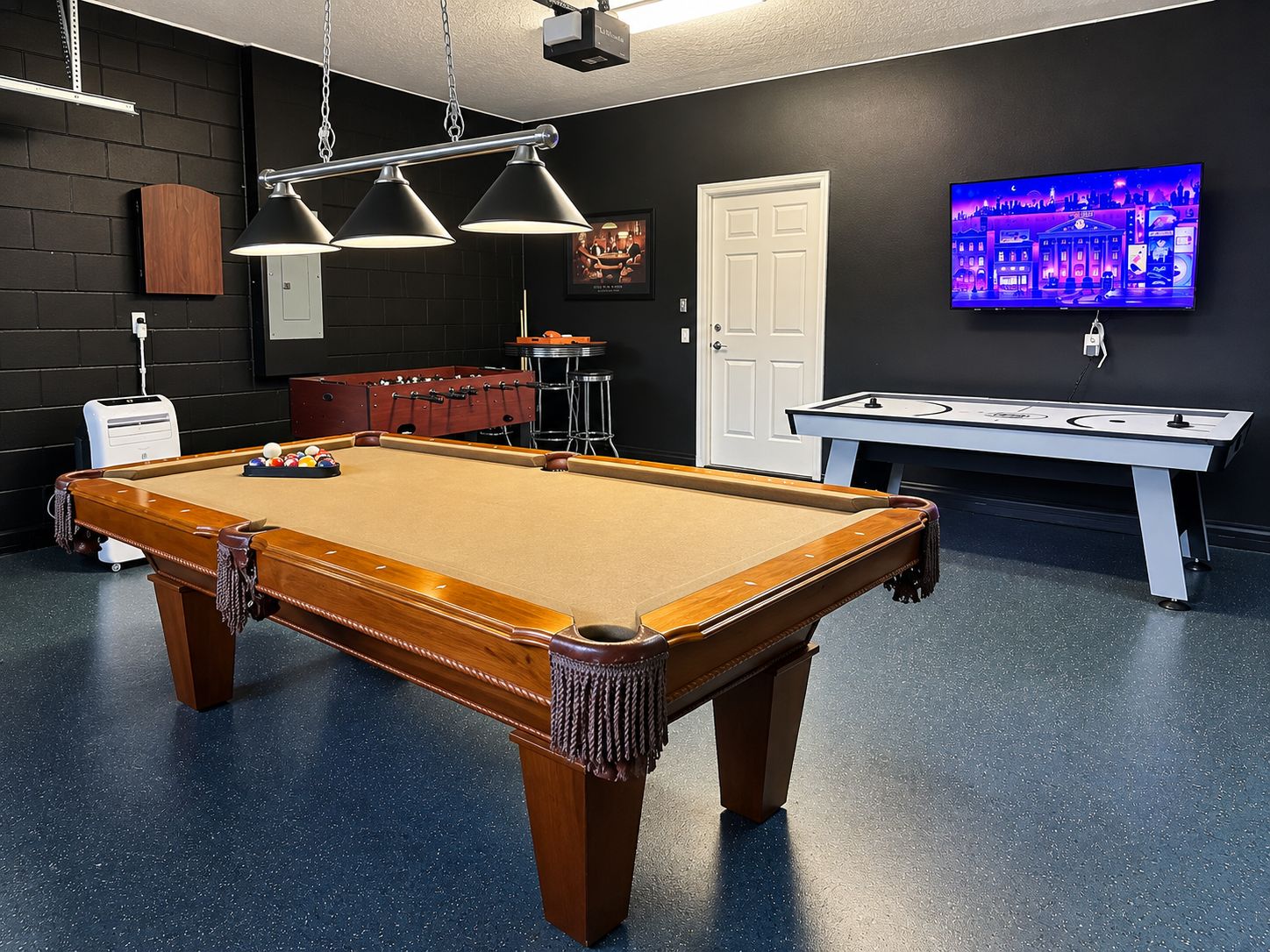 Una mesa de billar junto a una mesa de fútbol de mesa y un jukebox en una sala con pared oscura y televisor.