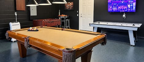 Una mesa de billar junto a una mesa de fútbol de mesa y un jukebox en una sala con pared oscura y televisor.