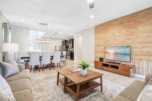 A modern living room with a wooden accent wall, a flat-screen TV, and a dining area with chairs and a table. - 6BR/4.5BA Kissimmee Home at Windsor at Westside, Pool (Kissimmee)