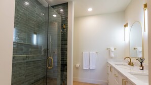 Een moderne badkamer met een douchecabine van glas, een wit wastafelmeubel met gouden handgrepen en een grote spiegel.